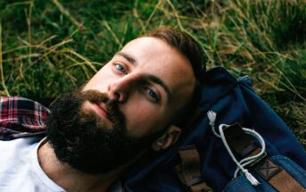 A bearded man lies on his back in the grass, resting his head on a blue backpack. He is wearing a plaid shirt over a white T-shirt and is looking directly at the camera.