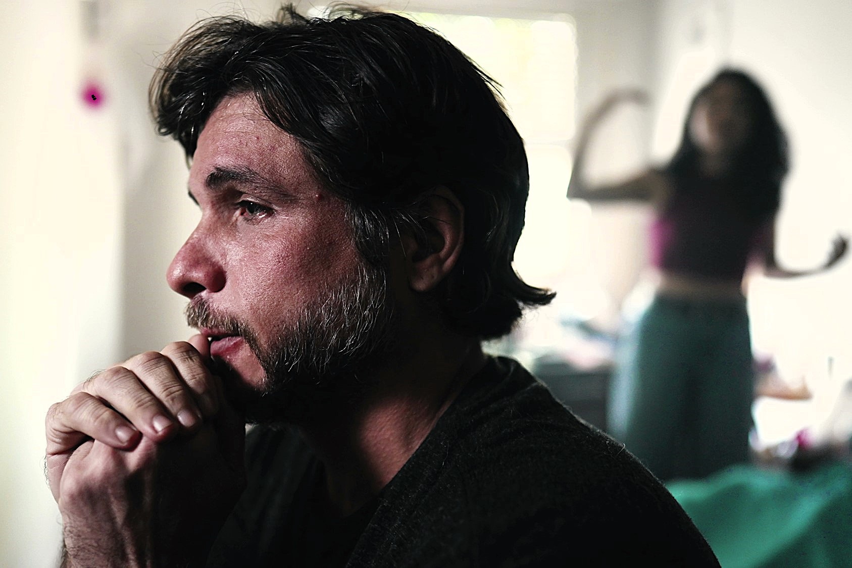 A man with dark hair and sideburns sits pensively with hands clasped under his chin, looking to the side. In the blurred background, a woman gestures with raised arms. The setting appears to be a dimly lit room.