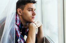 Young man in a plaid shirt leans on a windowsill, looking pensively outside. The soft lighting highlights his thoughtful expression as he rests his chin on his hands, framed by sheer curtains.