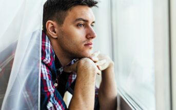 Young man in a plaid shirt leans on a windowsill, looking pensively outside. The soft lighting highlights his thoughtful expression as he rests his chin on his hands, framed by sheer curtains.