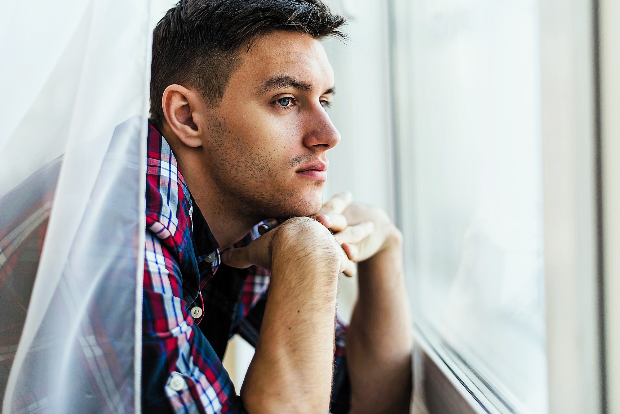 Young man in a plaid shirt leans on a windowsill, looking pensively outside. The soft lighting highlights his thoughtful expression as he rests his chin on his hands, framed by sheer curtains.