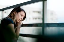 A woman leans on a balcony railing, looking pensively into the distance. She rests her chin on her hands, with a thoughtful expression. Buildings are blurred in the background.