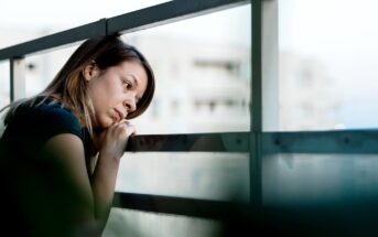 A woman leans on a balcony railing, looking pensively into the distance. She rests her chin on her hands, with a thoughtful expression. Buildings are blurred in the background.