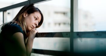 A woman leans on a balcony railing, looking pensively into the distance. She rests her chin on her hands, with a thoughtful expression. Buildings are blurred in the background.