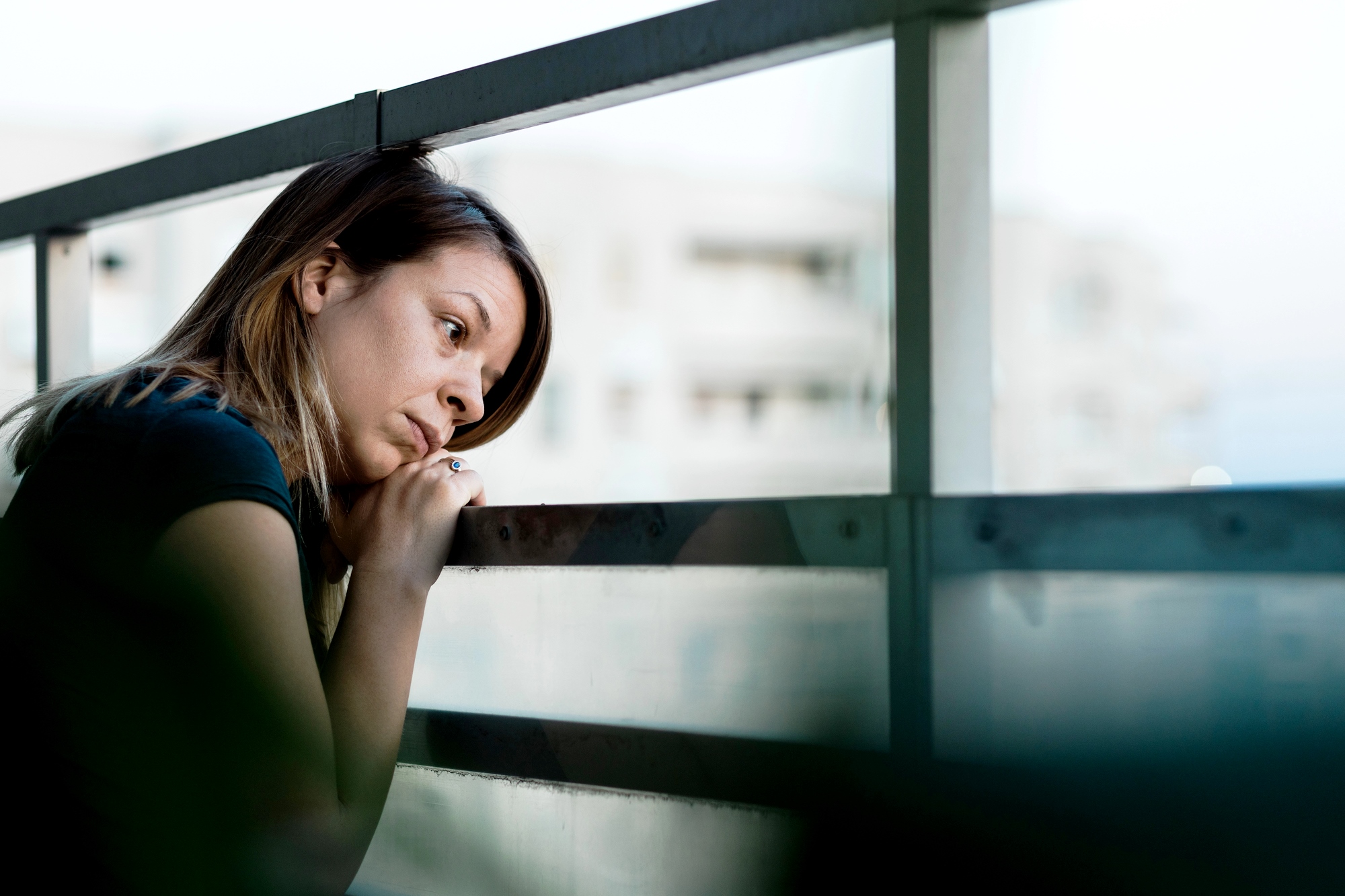 A woman leans on a balcony railing, looking pensively into the distance. She rests her chin on her hands, with a thoughtful expression. Buildings are blurred in the background.