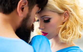 A close-up of a bearded man and a woman with blonde hair and bold makeup, touching foreheads. Both are wearing blue. The woman's eyes are closed, and they appear deeply connected.