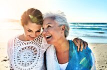 Two women smiling and embracing on a sunny beach. The woman on the left is wearing a white crocheted top, and the woman on the right is wearing a blue shawl. The ocean waves and bright sun create a warm, happy atmosphere.