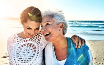 Two women smiling and embracing on a sunny beach. The woman on the left is wearing a white crocheted top, and the woman on the right is wearing a blue shawl. The ocean waves and bright sun create a warm, happy atmosphere.