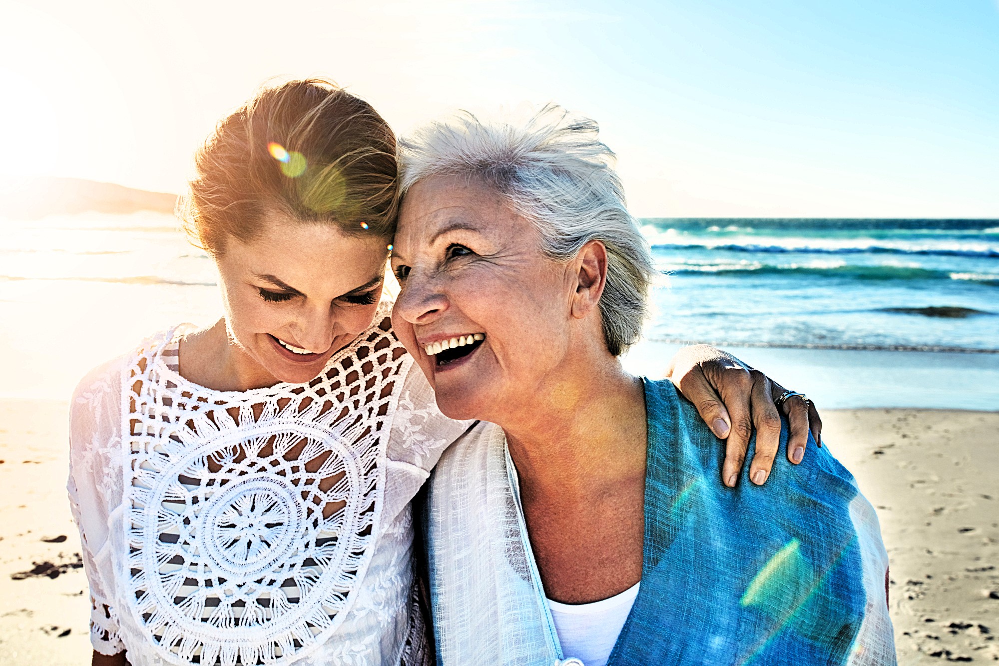 Two women smiling and embracing on a sunny beach. The woman on the left is wearing a white crocheted top, and the woman on the right is wearing a blue shawl. The ocean waves and bright sun create a warm, happy atmosphere.