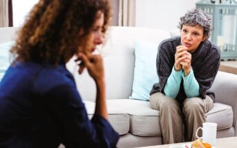 Two women sit on a beige sofa, engaged in conversation. One woman with curly hair listens intently, while the other with short gray hair speaks, leaning forward. Two white mugs and a plate with a pastry are on the table in front of them.