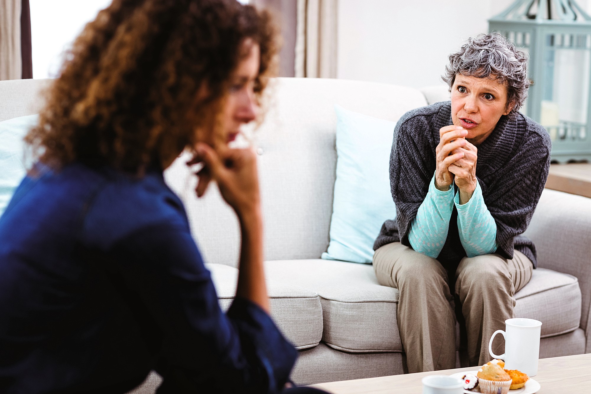 Two women sit on a beige sofa, engaged in conversation. One woman with curly hair listens intently, while the other with short gray hair speaks, leaning forward. Two white mugs and a plate with a pastry are on the table in front of them.