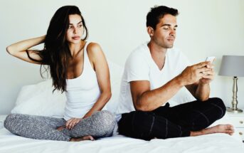 A woman and a man sit on a bed, both in casual clothing. The woman is looking away with her hand in her hair, while the man is focused on his smartphone. The room features white bedding and a bedside lamp.