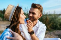 A man in a white shirt, wearing a watch, smiles as he holds a dark berry near a woman's lips. The woman, with long hair and a straw hat, smiles with closed eyes. They are outdoors with a blurred natural background under a clear blue sky.