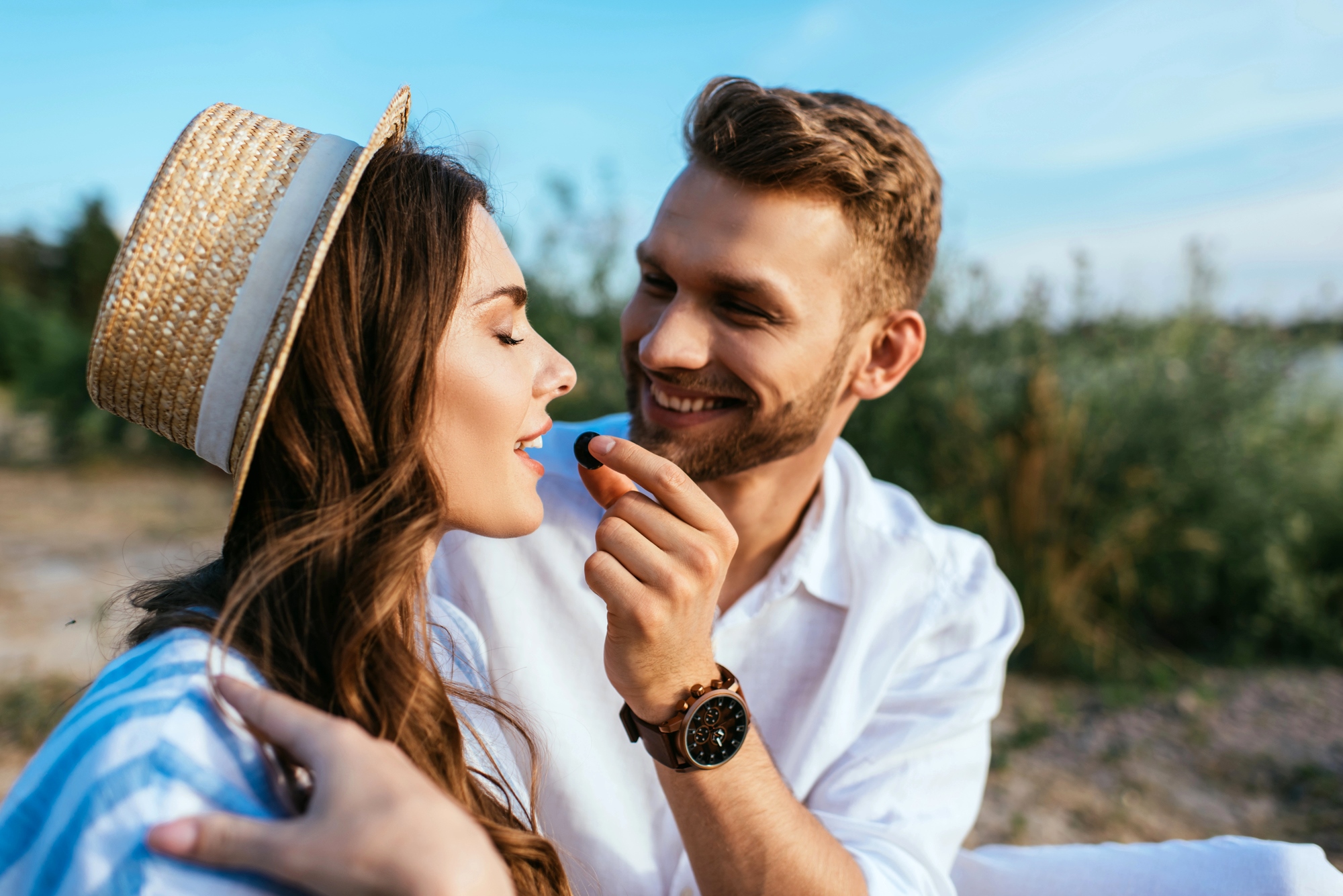 A man in a white shirt, wearing a watch, smiles as he holds a dark berry near a woman's lips. The woman, with long hair and a straw hat, smiles with closed eyes. They are outdoors with a blurred natural background under a clear blue sky.