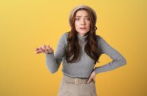 Woman with long brown hair wearing a beige hat, gray turtleneck, and beige pants. She stands against a yellow background, gesturing with one hand extended and the other on her hip, appearing confused or questioning.