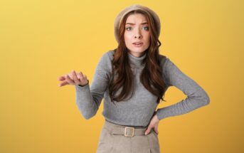 Woman with long brown hair wearing a beige hat, gray turtleneck, and beige pants. She stands against a yellow background, gesturing with one hand extended and the other on her hip, appearing confused or questioning.