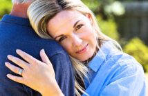 A woman with blonde hair smiles softly while resting her head on a man's shoulder. She is wearing a blue shirt and a ring on her left hand, embracing the man, who is wearing a dark blue shirt. The background is a blurred greenery.