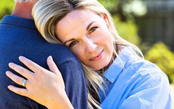 A woman with blonde hair smiles softly while resting her head on a man's shoulder. She is wearing a blue shirt and a ring on her left hand, embracing the man, who is wearing a dark blue shirt. The background is a blurred greenery.