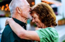 An elderly couple sharing an affectionate moment, the man gently kisses the woman's forehead while embracing her. They both smile warmly. String lights softly illuminate the background, creating a cozy atmosphere.