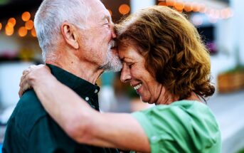 An elderly couple sharing an affectionate moment, the man gently kisses the woman's forehead while embracing her. They both smile warmly. String lights softly illuminate the background, creating a cozy atmosphere.