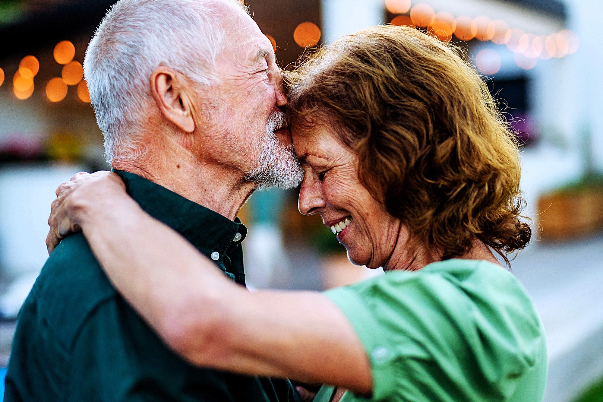 An elderly couple sharing an affectionate moment, the man gently kisses the woman's forehead while embracing her. They both smile warmly. String lights softly illuminate the background, creating a cozy atmosphere.