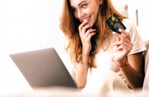 A smiling woman with long hair sits on a couch, holding a credit card in one hand and a laptop on her lap with the other. She appears excited or pleased, possibly online shopping or completing a purchase. The setting is bright and relaxed.