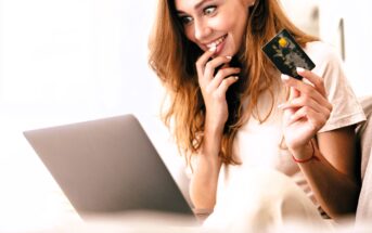 A smiling woman with long hair sits on a couch, holding a credit card in one hand and a laptop on her lap with the other. She appears excited or pleased, possibly online shopping or completing a purchase. The setting is bright and relaxed.