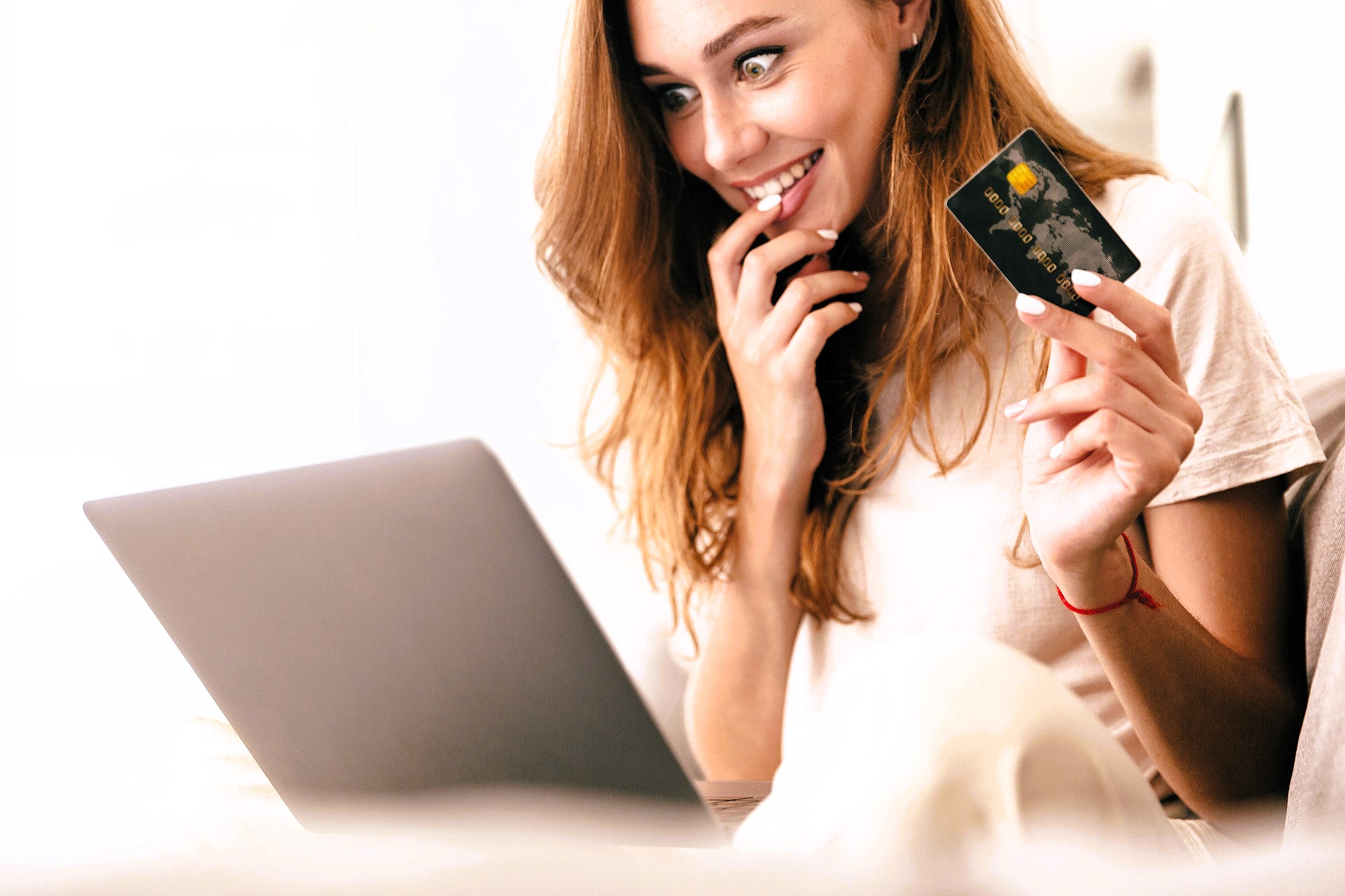 A smiling woman with long hair sits on a couch, holding a credit card in one hand and a laptop on her lap with the other. She appears excited or pleased, possibly online shopping or completing a purchase. The setting is bright and relaxed.