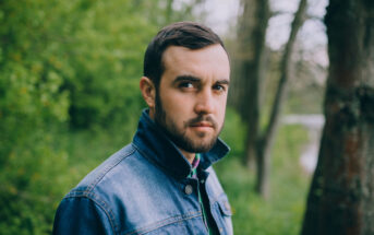 A man with short dark hair and a beard wears a denim jacket in an outdoor setting with green trees in the background. He looks intently at the camera.