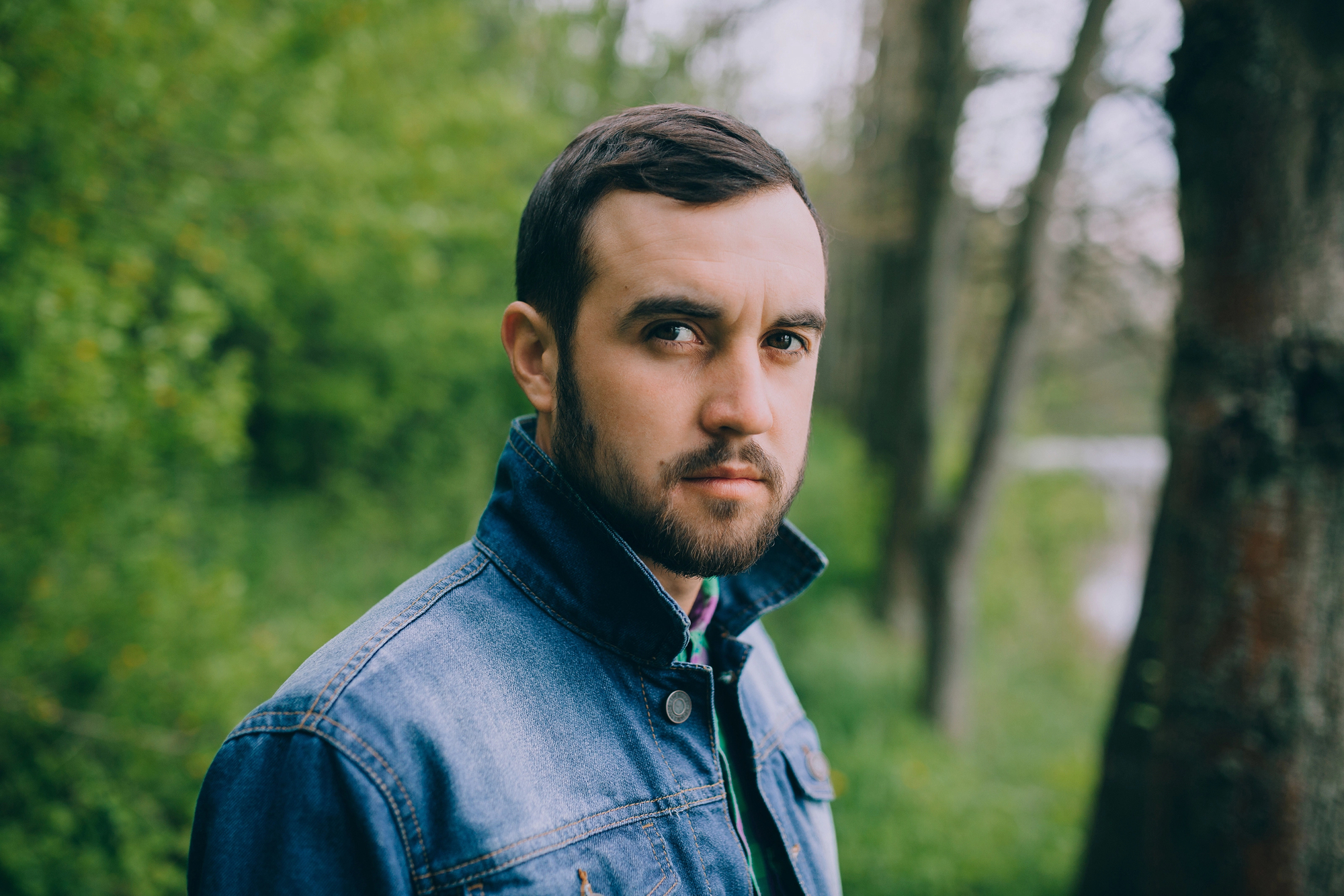 A man with short dark hair and a beard wears a denim jacket in an outdoor setting with green trees in the background. He looks intently at the camera.