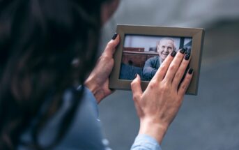 A person with dark hair and painted nails holds a framed photo of an older person smiling. The photo is being gently touched, suggesting a moment of reminiscence or affection.