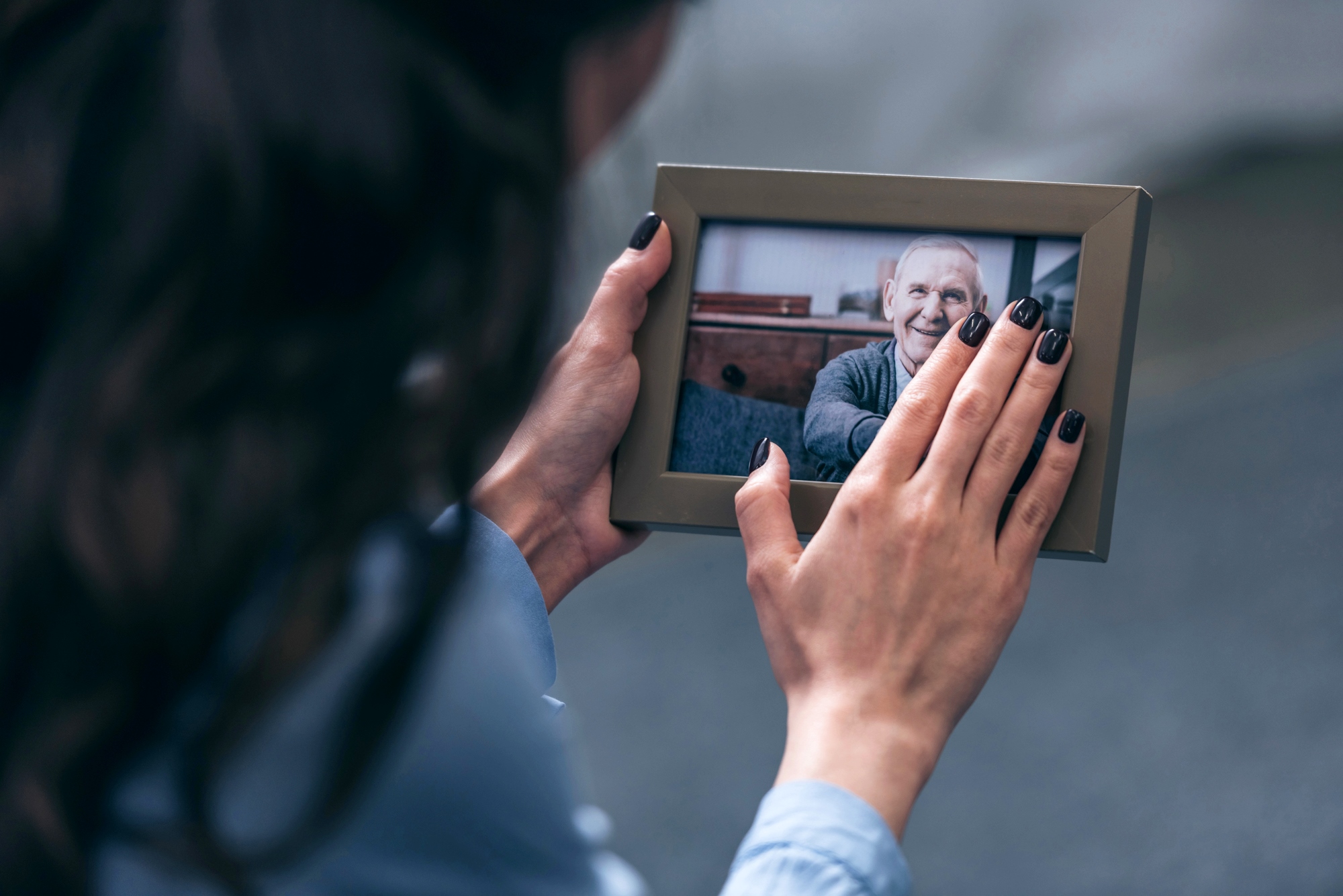 A person with dark hair and painted nails holds a framed photo of an older person smiling. The photo is being gently touched, suggesting a moment of reminiscence or affection.