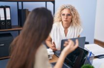 Two women engaged in a discussion at an office desk. The woman facing forward has curly hair and glasses, and the other has long hair, seen from behind. Laptops and office supplies are on the desk, with shelves and files in the background.