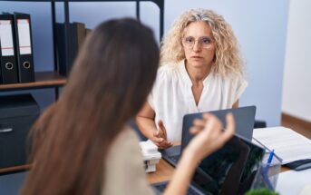 Two women engaged in a discussion at an office desk. The woman facing forward has curly hair and glasses, and the other has long hair, seen from behind. Laptops and office supplies are on the desk, with shelves and files in the background.