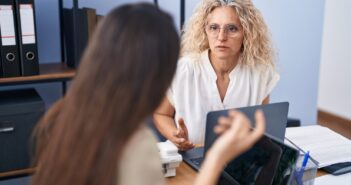 Two women engaged in a discussion at an office desk. The woman facing forward has curly hair and glasses, and the other has long hair, seen from behind. Laptops and office supplies are on the desk, with shelves and files in the background.