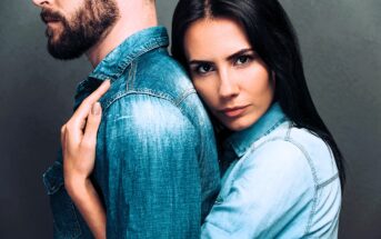 A woman with long dark hair embraces a bearded man from behind. Both are wearing denim shirts. The man faces away, while the woman looks directly at the camera with a serious expression. The background is a muted gray.