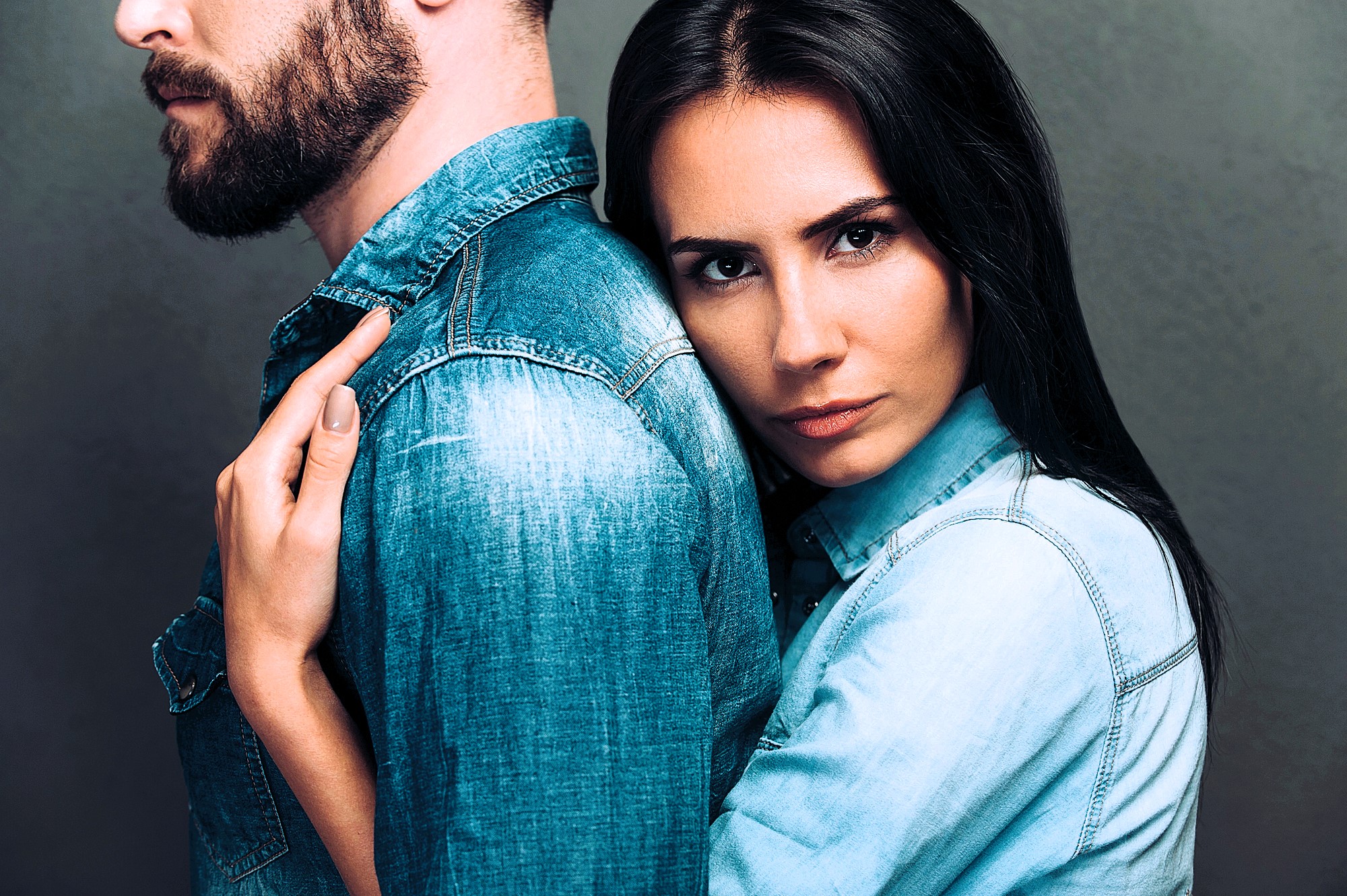 A woman with long dark hair embraces a bearded man from behind. Both are wearing denim shirts. The man faces away, while the woman looks directly at the camera with a serious expression. The background is a muted gray.