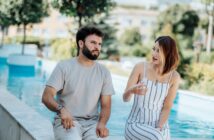 A man and a woman are sitting on a ledge beside a pool, engaged in a conversation. The man wears a light gray shirt and pants, while the woman is in a white and striped dress. Greenery and a blurred background are visible behind them.