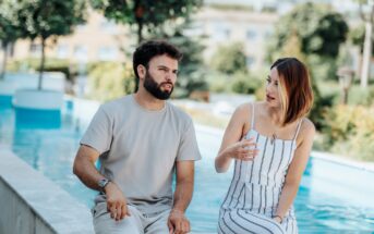 A man and a woman are sitting on a ledge beside a pool, engaged in a conversation. The man wears a light gray shirt and pants, while the woman is in a white and striped dress. Greenery and a blurred background are visible behind them.