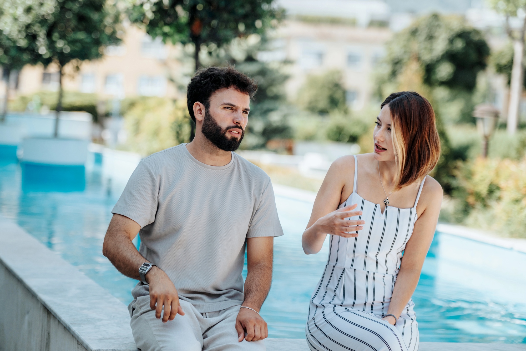 A man and a woman are sitting on a ledge beside a pool, engaged in a conversation. The man wears a light gray shirt and pants, while the woman is in a white and striped dress. Greenery and a blurred background are visible behind them.