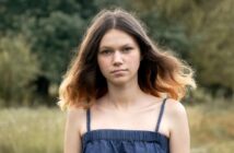 A young woman with long, wavy hair stands outdoors, looking at the camera. She is wearing a blue sleeveless top. The background is blurred with greenery, suggesting a natural setting.