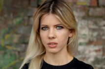 A person with long blonde hair and blue eyes is wearing a black shirt and standing against a blurred brick wall background. They have a neutral expression.