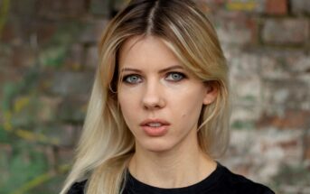 A person with long blonde hair and blue eyes is wearing a black shirt and standing against a blurred brick wall background. They have a neutral expression.