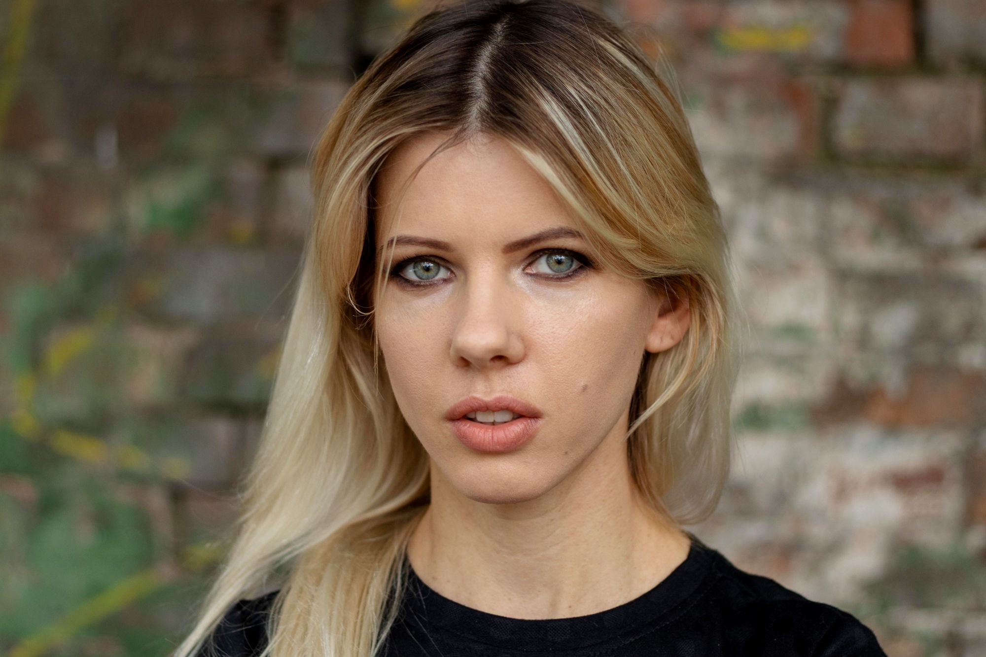 A person with long blonde hair and blue eyes is wearing a black shirt and standing against a blurred brick wall background. They have a neutral expression.