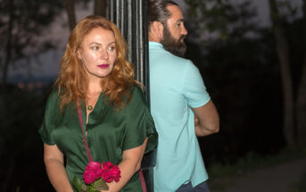 A woman with red hair holds pink roses, looking away pensively. She stands beside a lamppost. A bearded man in a light blue shirt stands on the other side, facing away. They are outdoors in a dimly lit setting.