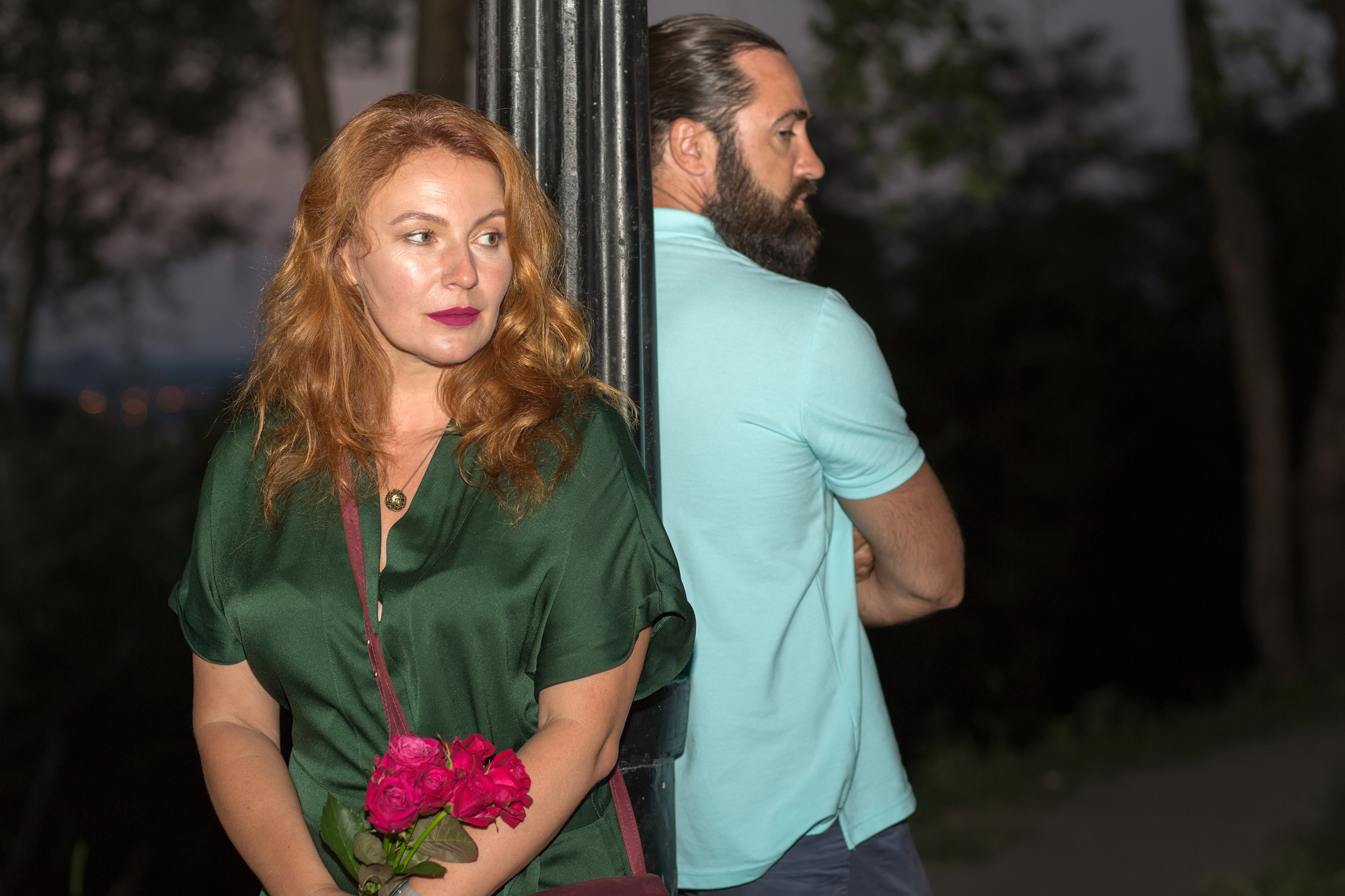 A woman with red hair holds pink roses, looking away pensively. She stands beside a lamppost. A bearded man in a light blue shirt stands on the other side, facing away. They are outdoors in a dimly lit setting.