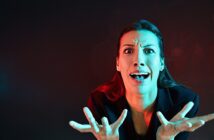 A woman with long hair is expressing shock or frustration. Her hands are raised in front of her with fingers spread wide. The lighting is dramatic, with red and blue tones, set against a dark background.