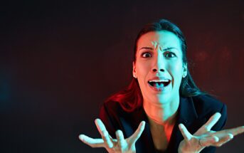 A woman with long hair is expressing shock or frustration. Her hands are raised in front of her with fingers spread wide. The lighting is dramatic, with red and blue tones, set against a dark background.