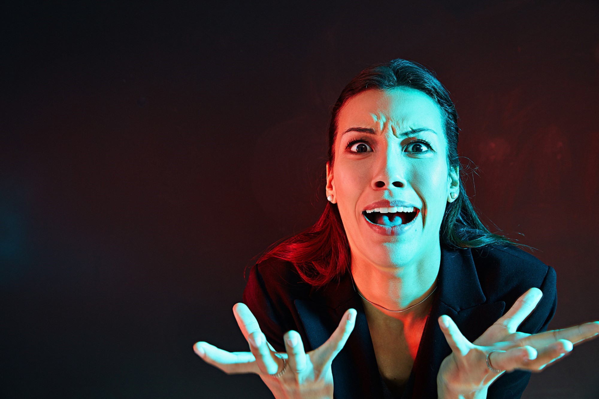 A woman with long hair is expressing shock or frustration. Her hands are raised in front of her with fingers spread wide. The lighting is dramatic, with red and blue tones, set against a dark background.