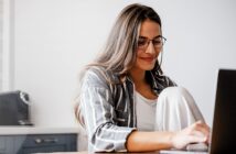 A woman with long hair and glasses is sitting at a table, smiling while working on a laptop. She is wearing a striped shirt over a white top, and appears relaxed in a casual home setting.